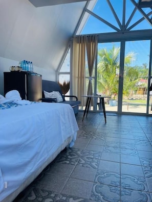 Een hotelkamer met een bed, een kleintje tafeltje, een stoel, een minibar en uitzicht op palmbomen door grote glazen deuren.