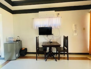 Een kleine kamer met een tafel en stoelen, een aan de muur bevestigde televisie en een koelkast.