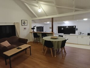 A living room with a sofa, a coffee table, and a dining area with a table and chairs. There is a kitchen area with cabinets and a window. - Beautiful Byron hinterland retreat. 20 mins to Byron Bay, Bangalow and Ballina. (Clunes)