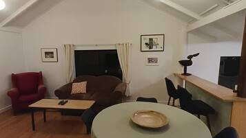 Uma sala de estar acolhedora com um sofá marrom, uma poltrona vermelha, uma mesa pequena e um espaço de jantar com uma mesa redonda e cadeiras.