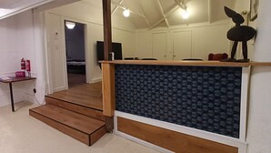 Una habitación con escalera de madera, una pequeña mesa con extintor, una zona de kitchenette y techo con vigas a la vista.