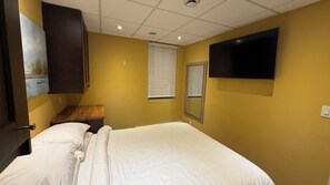 Une chambre d’hôtel avec un lit, un téléviseur fixé au mur, une fenêtre avec des stores et un tableau accroché au mur.