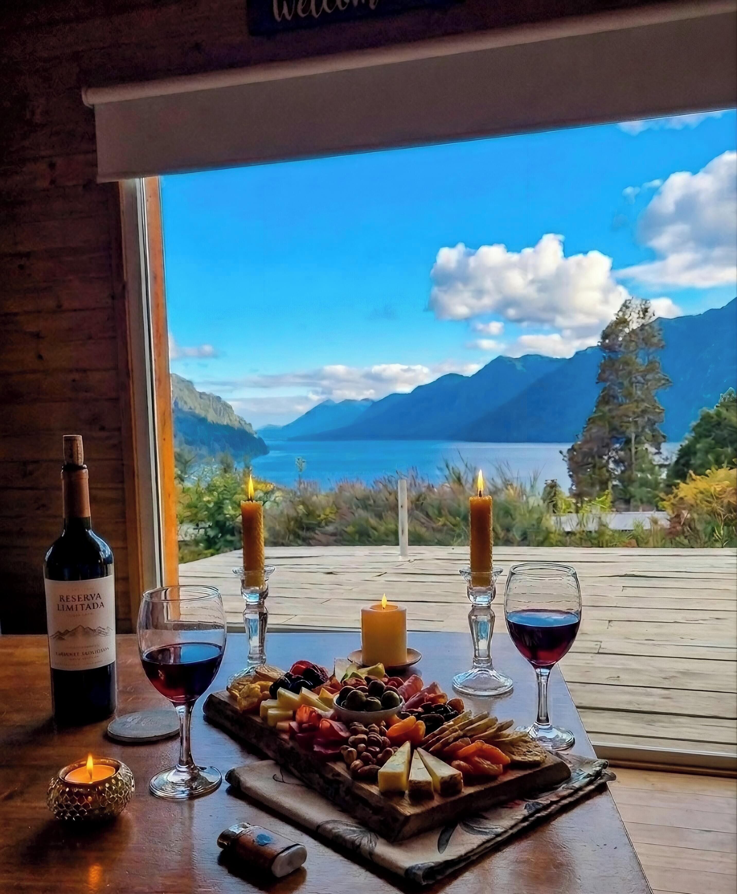 Una mesa con una botella de vino, copas, una vela y una bandeja de frutas junto a una ventana con vistas panorámicas.