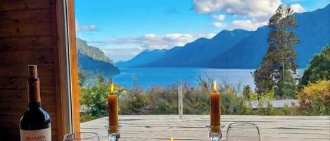 Una mesa con una botella de vino, copas, una vela y una bandeja de frutas junto a una ventana con vistas panorámicas.