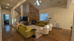 A spacious living room with a stone fireplace, a staircase, and a large TV. - Upper River Cottage - Main Floor & Upstairs, 3-beds, near SF River trail & parks (Spanish Fork)