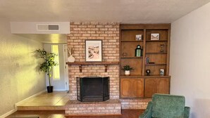 A living room with a brick fireplace, a green armchair, a wooden bookshelf, and a potted plant. - El Paso | Large Group + RV Parking (El Paso)