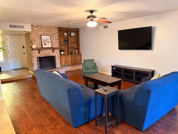 A living room with a brick fireplace, blue sofas, a television, and a ceiling fan.