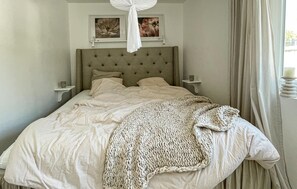 Une chambre à coucher avec une tête de lit capitonnée, deux tables de chevet, un lit recouvert d’une couverture tricotée et deux tableaux encadrés accrochés au mur.