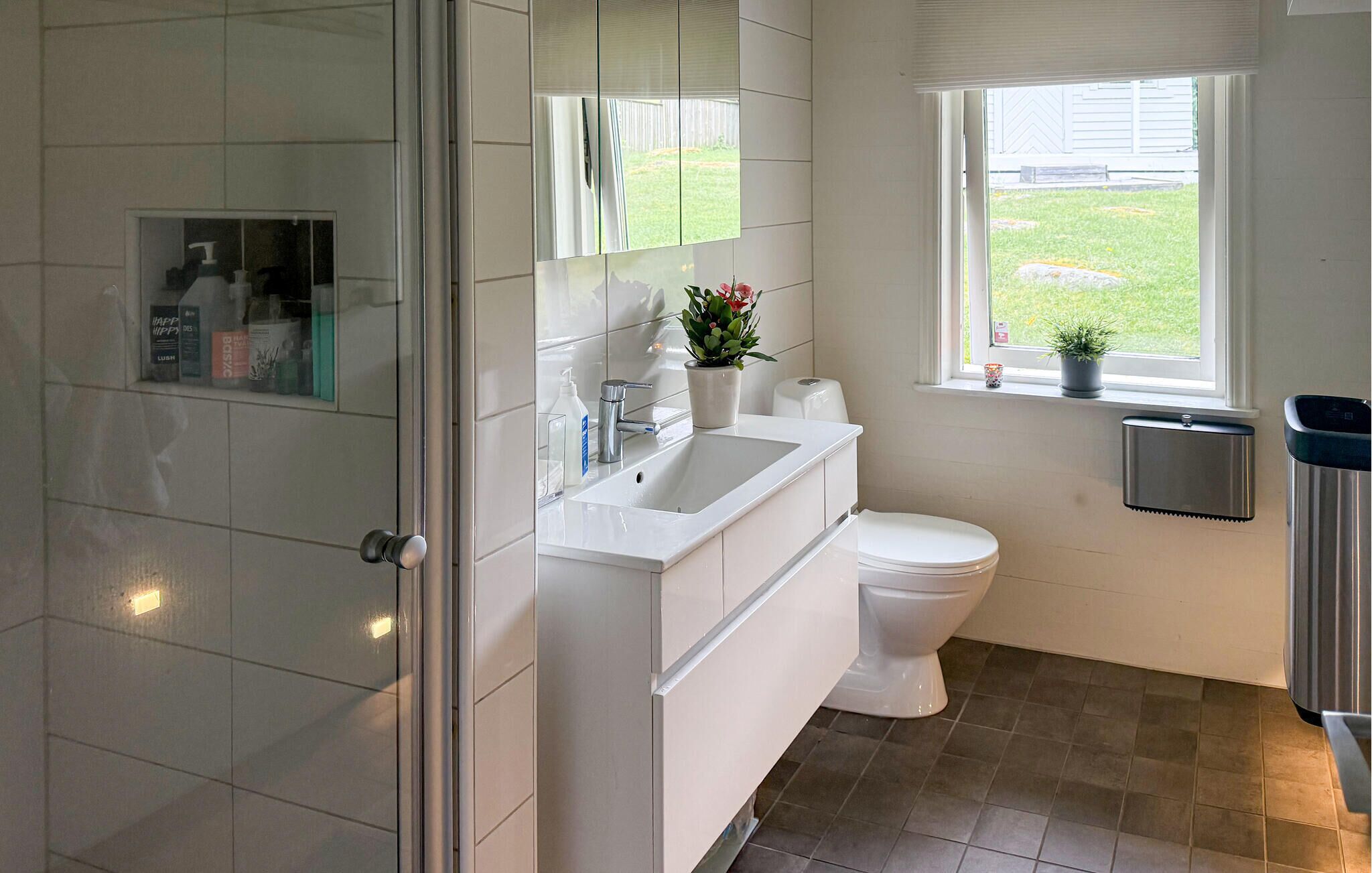 Un bany modern amb una cabina de dutxa de vidre, un lavabo blanc, un vàter i una finestra amb vista al jardí.
