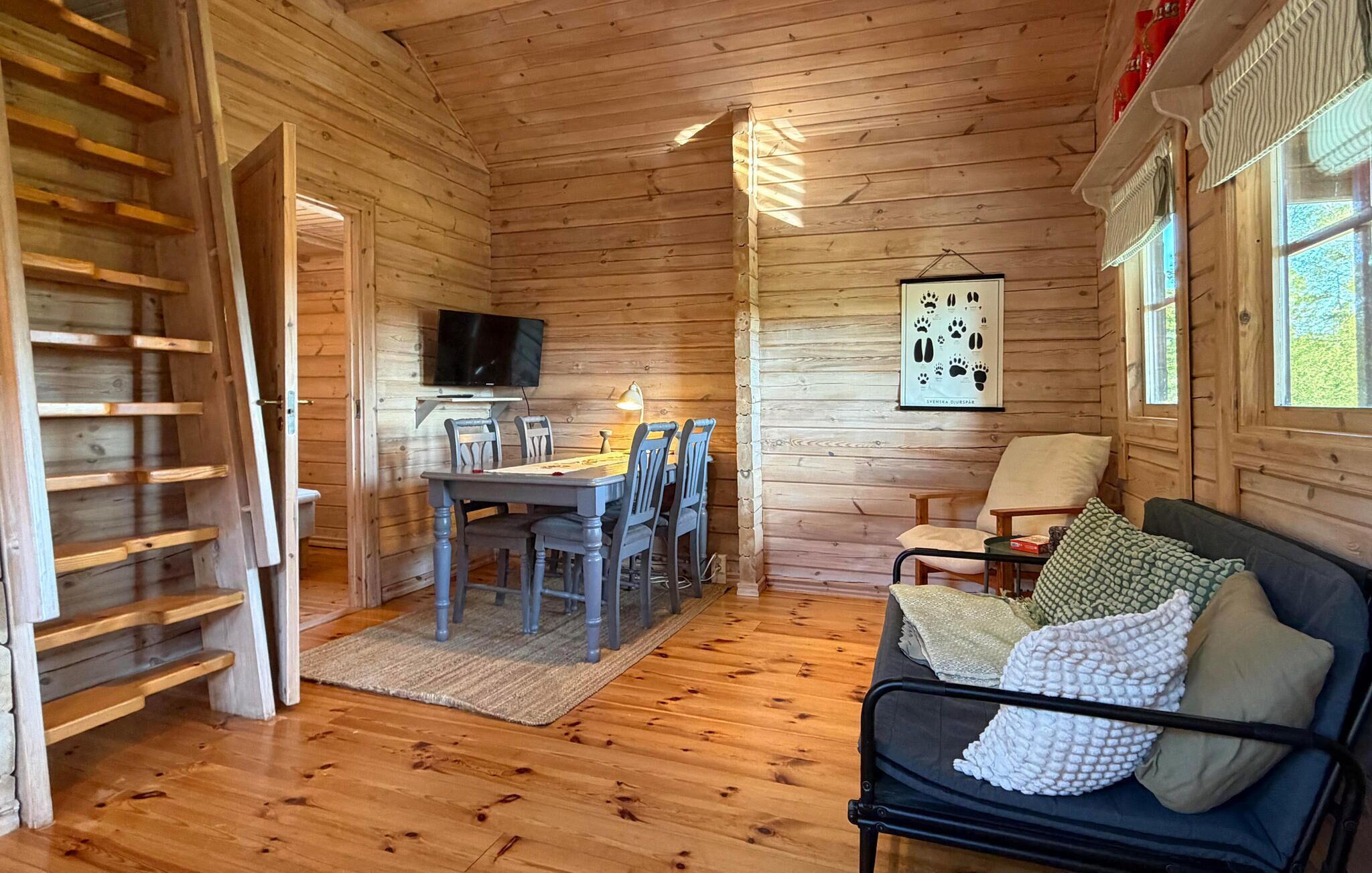 Een houten chalet van binnen met een eettafel, stoelen, een bank, een televisie en een poster aan de muur.
