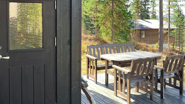 Een houten terras met een tafel, stoelen, een deur en een blokhut op de achtergrond.