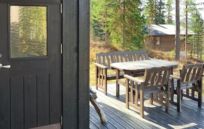 A wooden deck with a table and chairs, a door, and a cabin in the background.