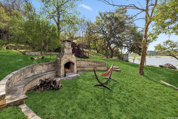 A stone fire pit with a chimney, surrounded by a stone wall, in a grassy area with trees and a view of a body of water. - Charming Waterfront Cabin on Lake Travis (Leander)