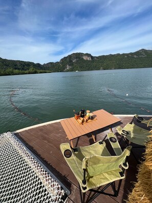En træterrasse med et spisebord, omgivet af grønne stole, med udsigt over en rolig sø med både og grønt i baggrunden.