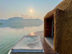 A thatched-roof structure with a wooden door, a woven mat on the deck, and a view of calm water and distant mountains.