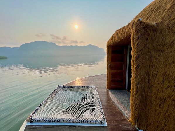 A thatched-roof structure with a wooden door, a woven mat on the deck, and a view of calm water and distant mountains.