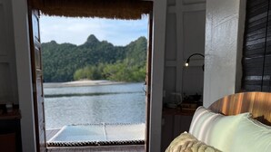 Un dormitorio con una cama, una mesita de noche, una lámpara y vistas a un lago y montañas a través de una puerta abierta.