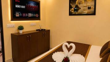 A hotel room with a bed, a TV displaying a Netflix series, a painting, and a cabinet with a plant.