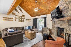 A cozy living room with a stone fireplace, wooden ceiling beams, and a large window. - Camelback Family Escape | Ski-In/Out + Game Room (Tannersville)