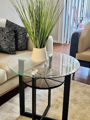 Una sala de estar con una mesa de centro de vidrio, una planta en maceta y cojines estampados.