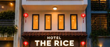 L’entrée d’un hôtel, avec une enseigne indiquant « HÔTEL THE RICE », un comptoir d’accueil et un escalier menant aux étages supérieurs.
