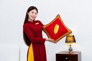 A person holding a red and yellow pillow with a gold emblem, standing next to a bedside table with a lamp. - The Rice Hotel - Ben Thanh (Ho Chi Minh City)