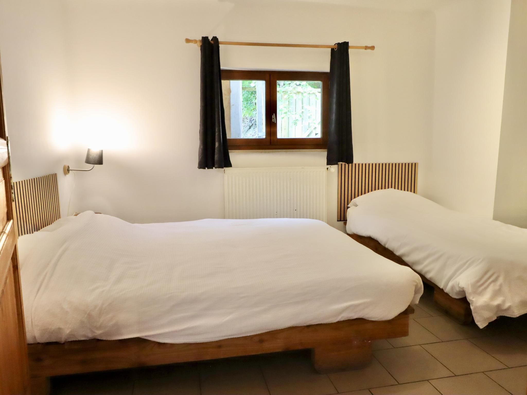 Une pièce avec deux lits en bois, une fenêtre avec des rideaux, un radiateur et une lampe fixée au mur.