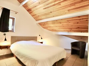 A bedroom with a sloped ceiling, a bed, bedside lamps, a window, and a chair. - Pretty group house with a sauna, games & garden (Lierneux)