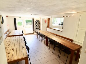 A dining area with wooden tables and chairs, a kitchen area with a window, and a door leading outside. - Pretty group house with a sauna, games & garden (Lierneux)