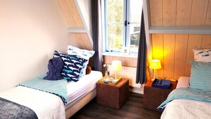 Ein Zimmer mit zwei Betten, einem Fenster mit Vorhängen, einer Lampe und einem kleinen Tisch.