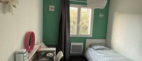 Um quarto pequeno com uma cama, uma escrivaninha com cadeira, uma janela com cortinas, um radiador e uma luminária suspensa.