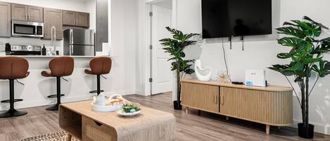 A modern kitchen with stainless steel appliances, a dining area with a wooden table and chairs, a flat-screen TV mounted on the wall, and a potted plant.
