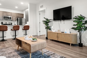 A modern kitchen with stainless steel appliances, a dining area with a wooden table and chairs, a flat-screen TV mounted on the wall, and a potted plant.