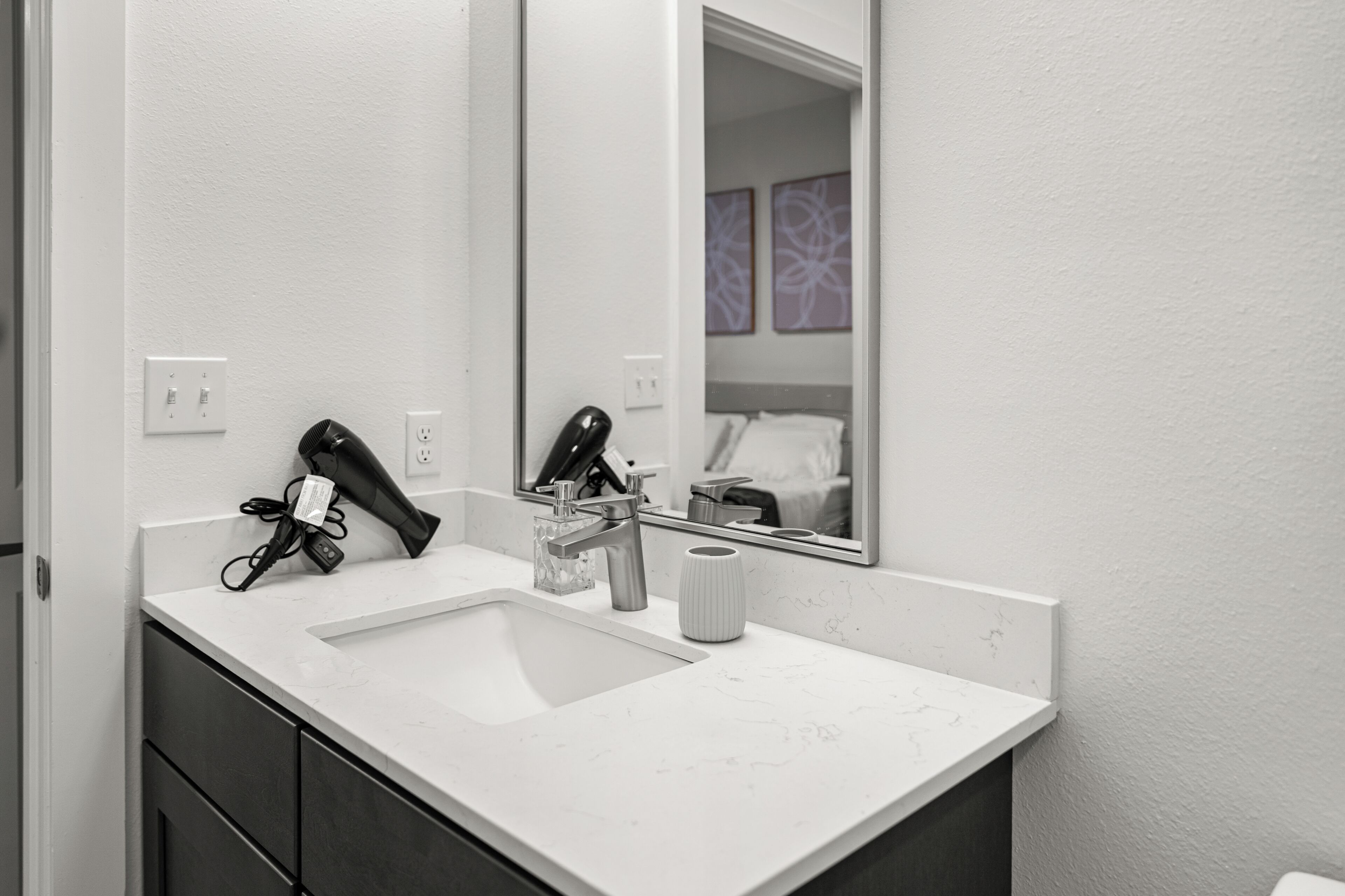 Un baño moderno con lavamanos blanco, un espejo, un secador de pelo y un perchero para toallas.