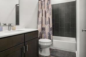 Une salle de bain avec des toilettes blanches, des murs recouverts de carreaux foncés, un rideau de douche et une baignoire.