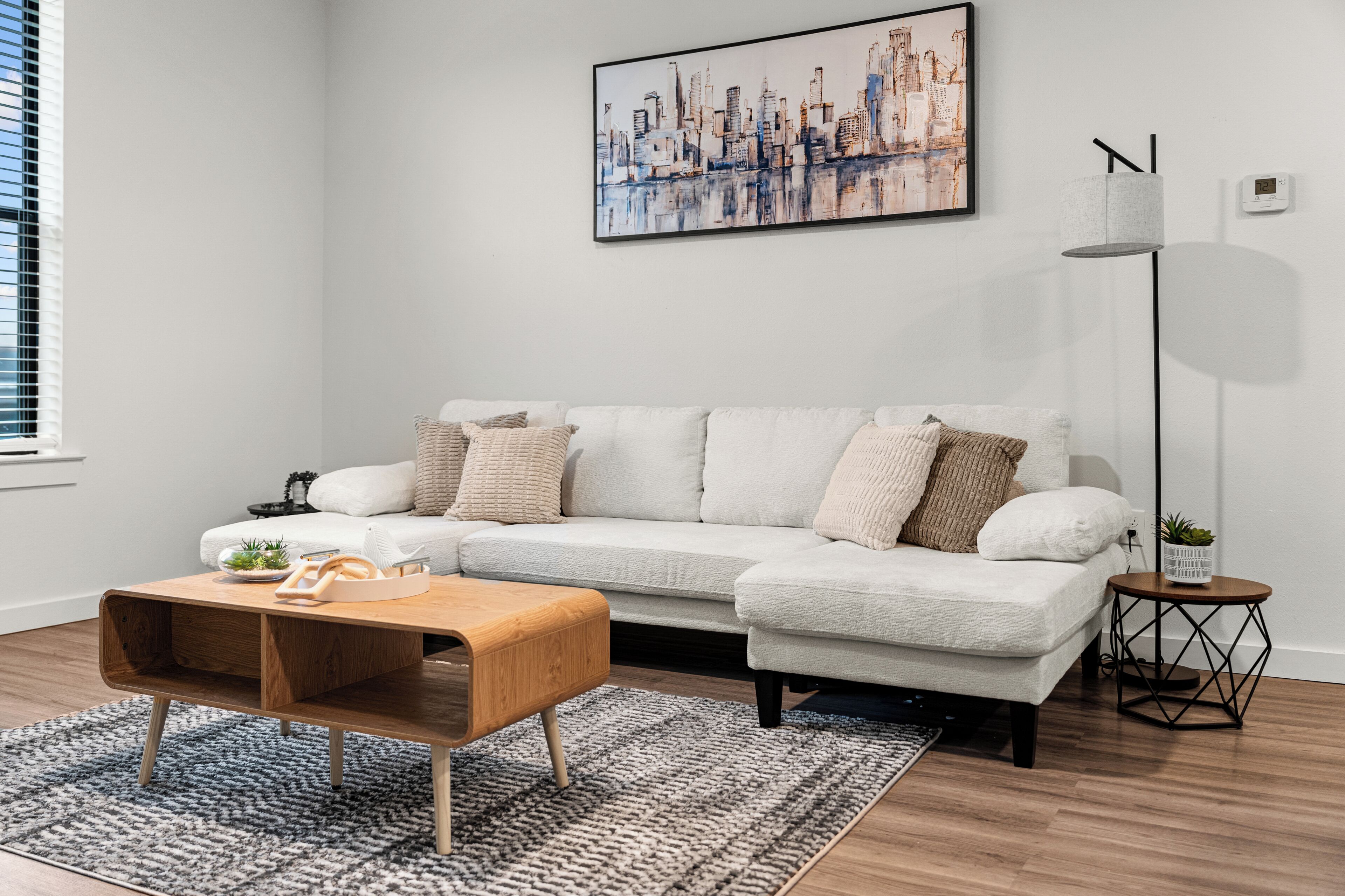 Una sala de estar moderna con un sofá blanco, una mesa de centro de madera, un cuadro de un paisaje urbano, una lámpara de pie y una pequeña mesita con una planta.