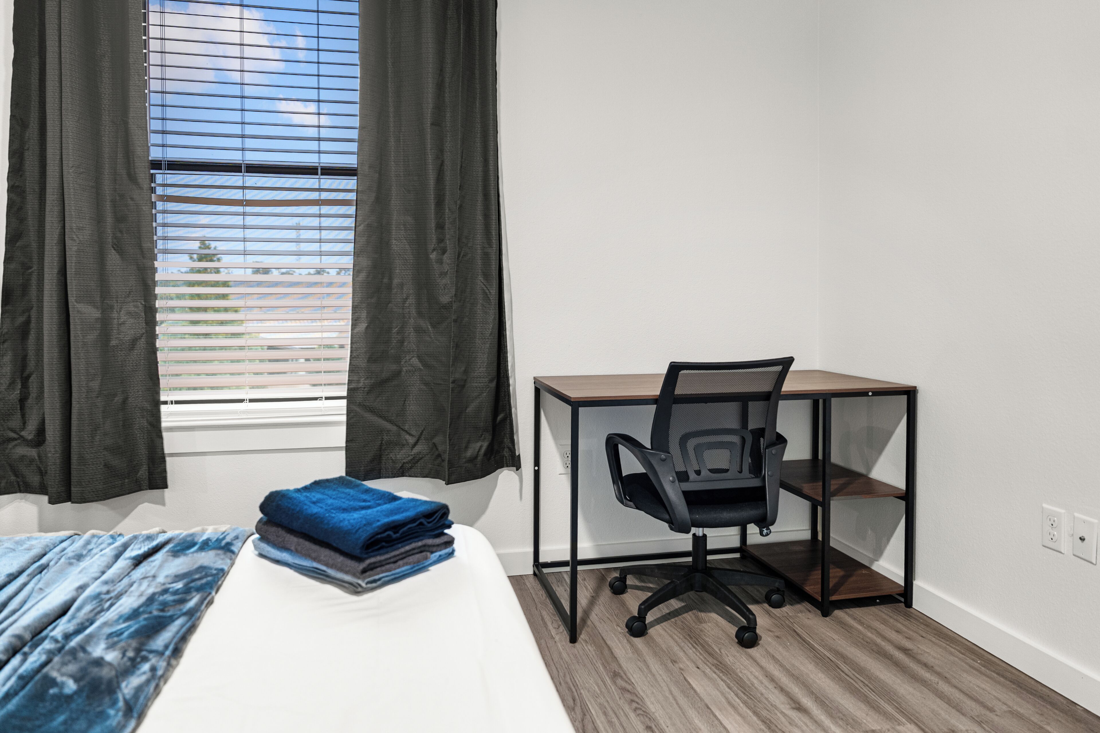 Una habitación con cama, escritorio con silla, ventana con cortinas y vista al exterior.