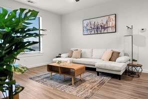 Un salon moderne avec un canapé blanc, une table basse en bois, un tableau représentant un paysage urbain et un lampadaire.