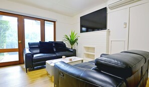 Uma sala de estar com um sofá de couro preto, uma mesa de centro branca e uma TV de tela plana fixada em uma parede de tijolos brancos.