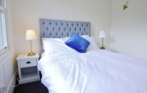 Una cama bien tendida con sábanas blancas y un cojín azul decorativo, una mesita de noche con lámpara, y un aplique en la pared.