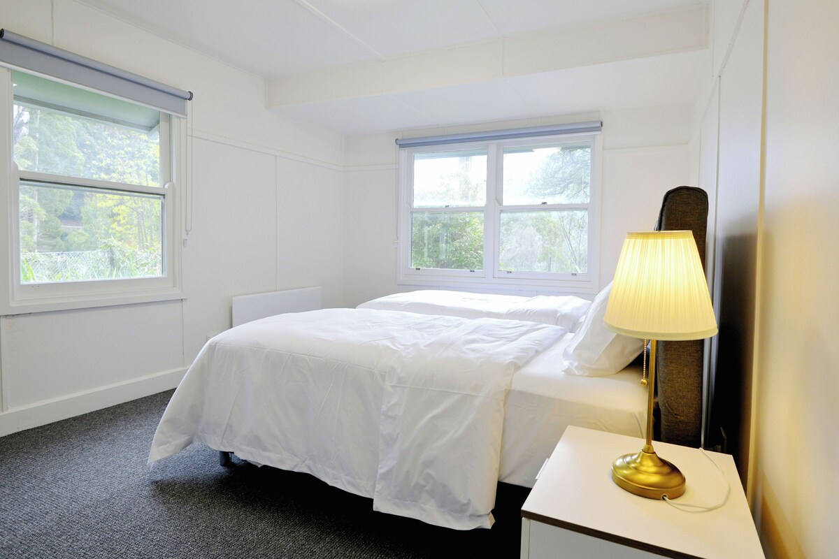 A hotel room with a bed, a bedside table with a lamp, and two windows.