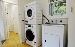 A laundry room with stacked washing machines, a dryer, and a window with a view of greenery.
