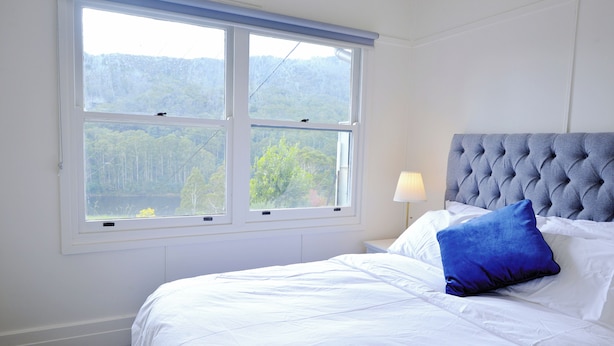 Une chambre avec une grande fenêtre, un lit avec une literie blanche, un oreiller bleu et une tête de lit grise à motifs.