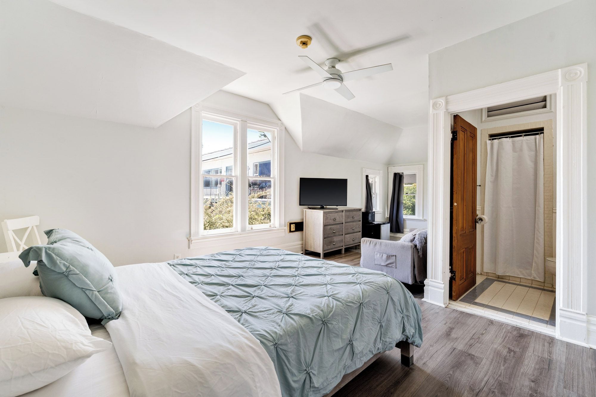 A bedroom with a bed, a television, a ceiling fan, and a view of the outside.