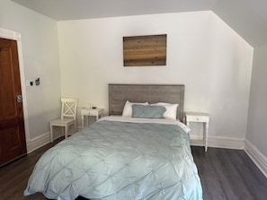 Een slaapkamer met een bed, een houten hoofdbord, een nachtkastje, een stoel en een kunstwerk aan de muur.