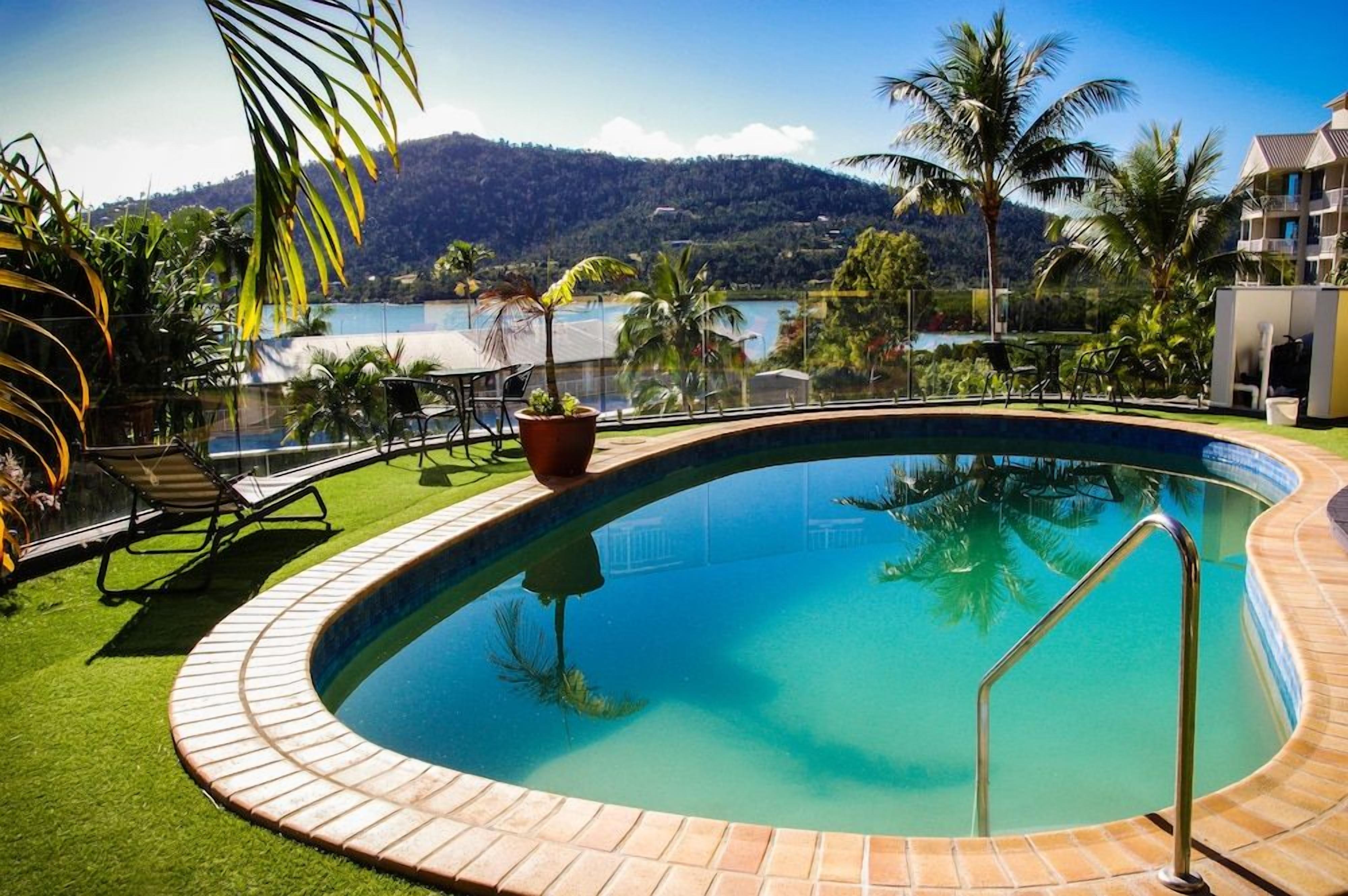 Piscina con agua azul cristalina, rodeada por un borde de ladrillo, y vista a una montaña y un lago al fondo.