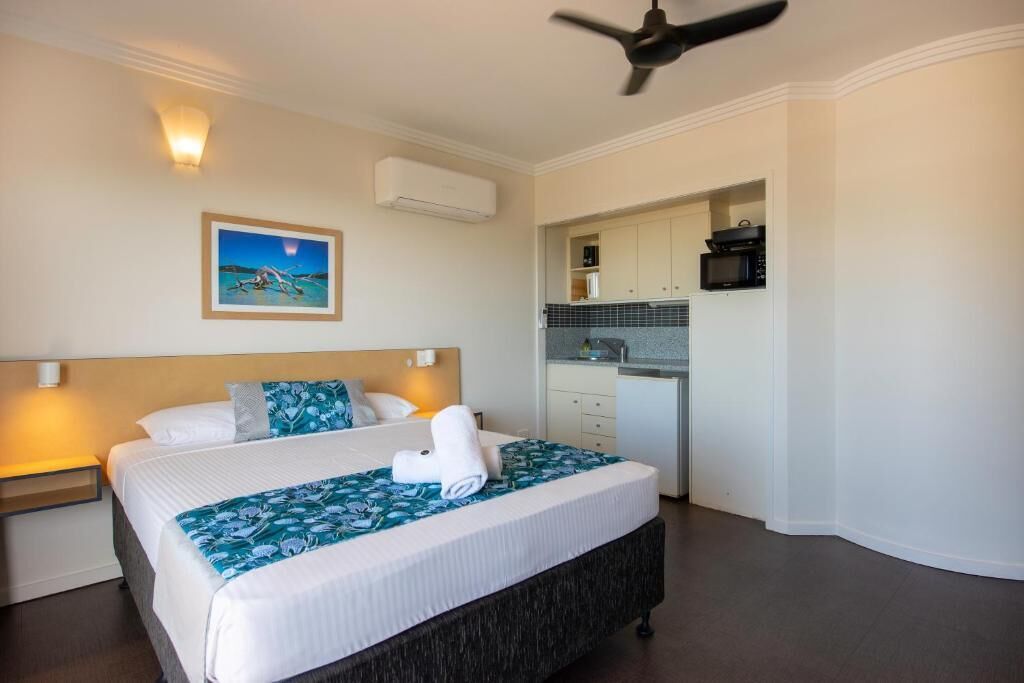 Una camera d'albergo con un letto, un comodino, un condizionatore d'aria a parete, una ventola a soffitto, un angolo cottura con pensili ed elettrodomestici, e un quadro incorniciato appeso al muro.