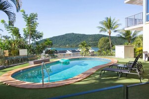 A swimming pool with a view of a lake and hills, surrounded by palm trees and a building with a balcony.
