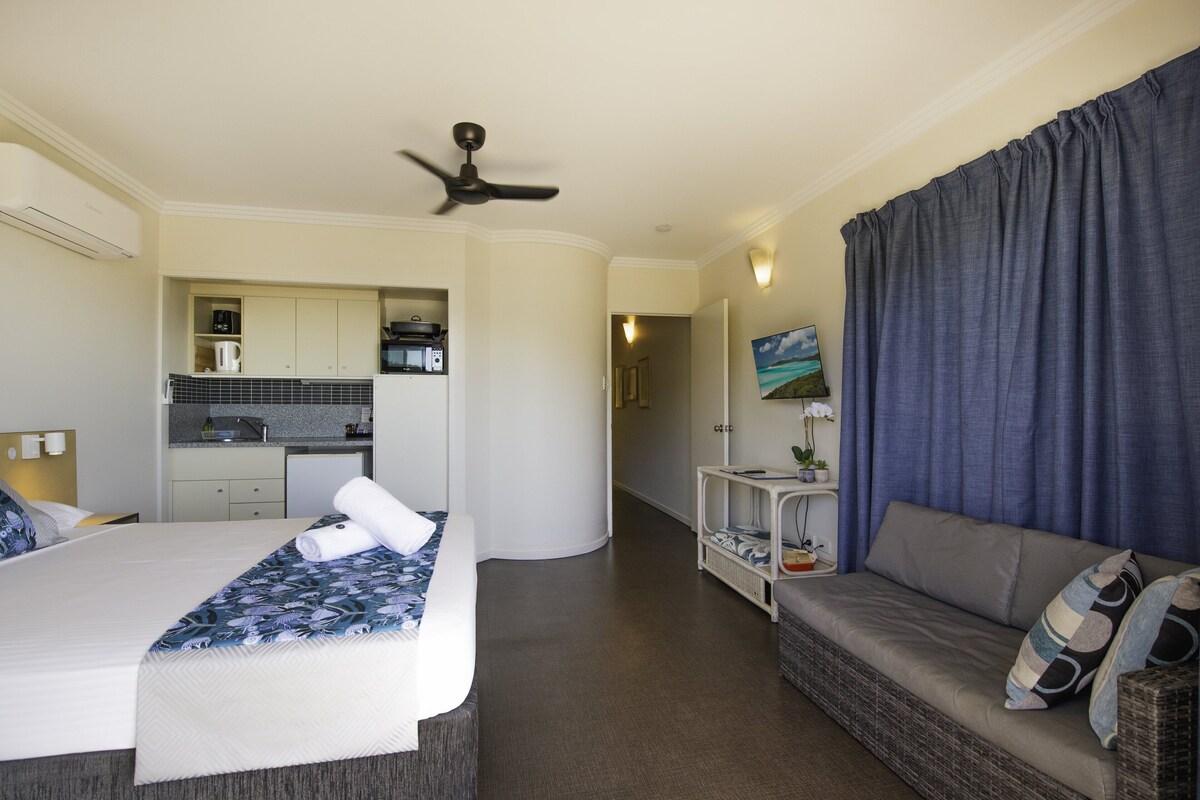 A hotel room with a bed, a sofa, a kitchenette, a ceiling fan, and a painting.