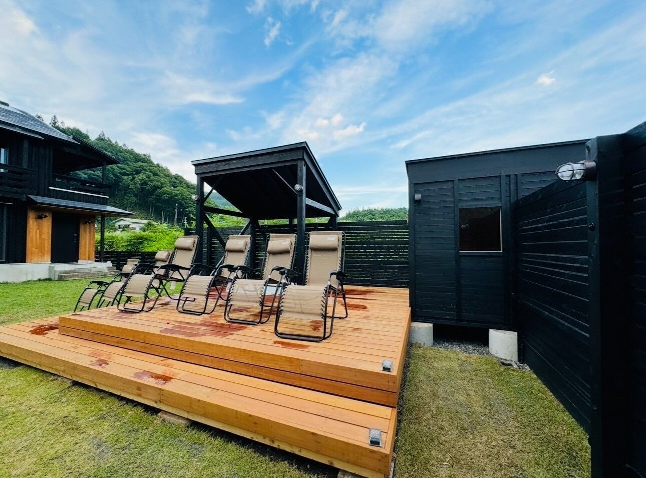 A deck with lounge chairs and a covered seating area, surrounded by a grassy lawn and a black building.